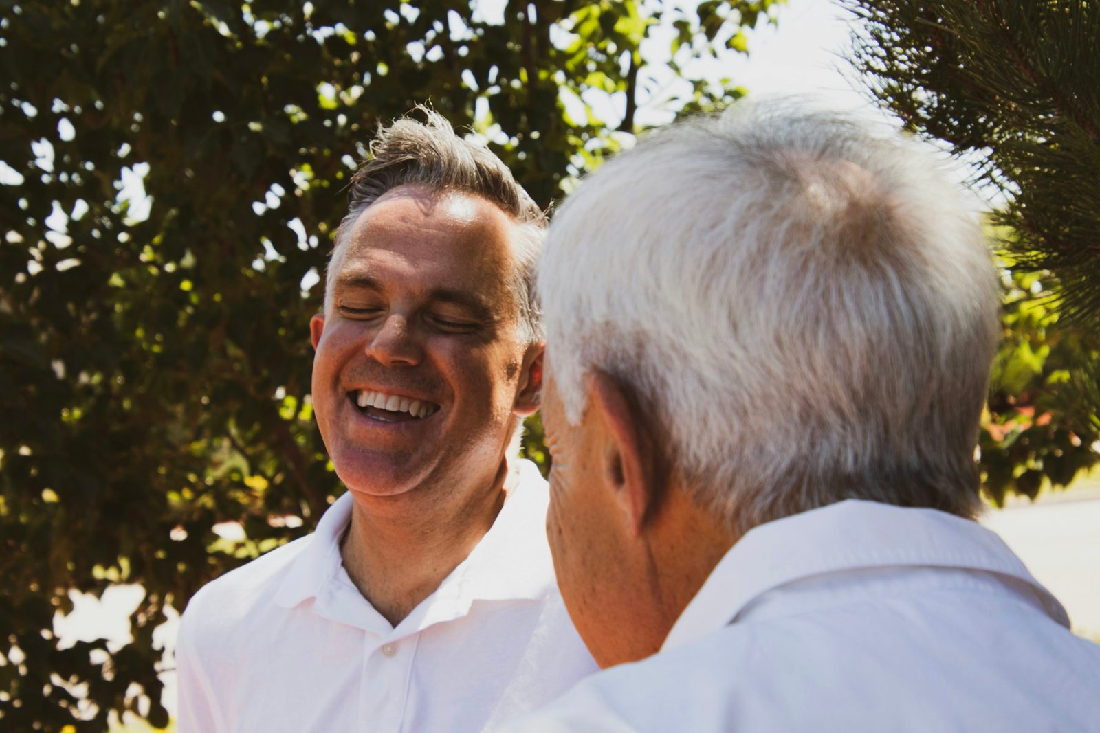 A younger and an older man sharing a laugh outdoors under trees