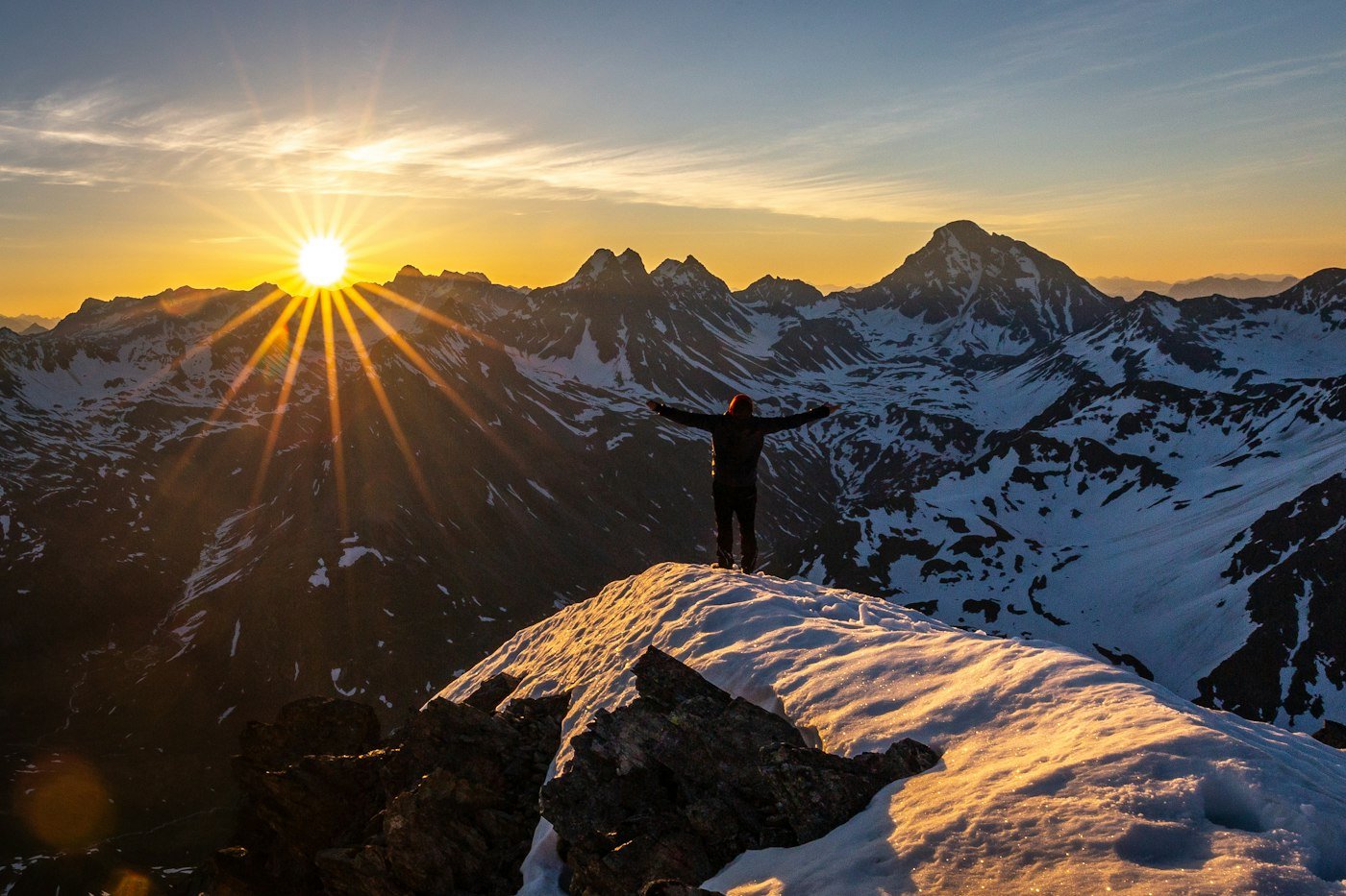 A person standing on a snowy rock at sunset in the mountains
