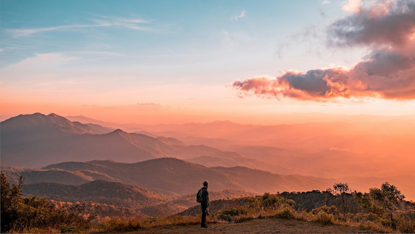 A man standing alone on a hillside looking out over layered mountains at sunset